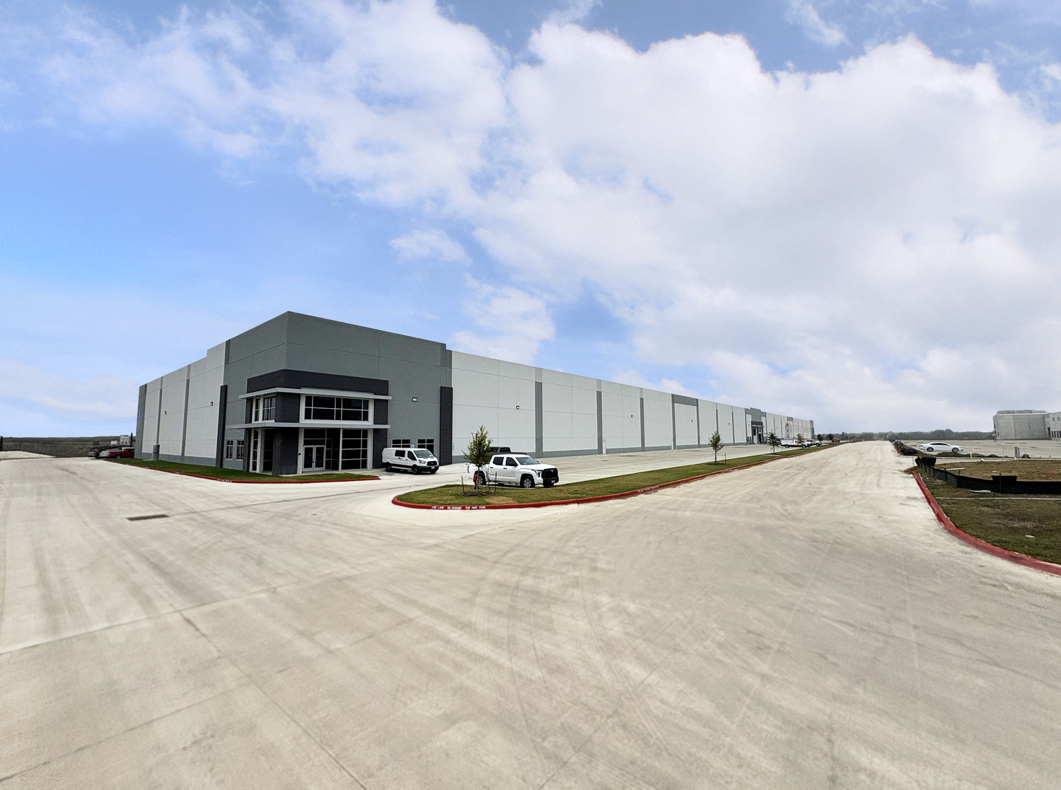 A large, modern warehouse building with white and gray walls sits at the corner of wide, empty paved roads. Several pickup trucks are parked near the entrance. The sky is partly cloudy.