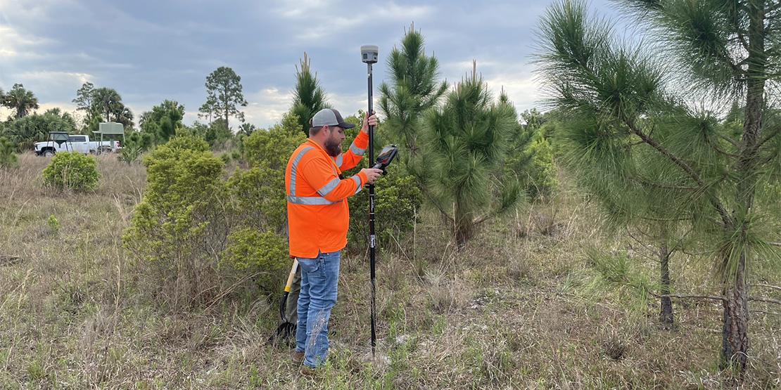 A BGE team member in an orange hazard vest setting up survey equipment in brush