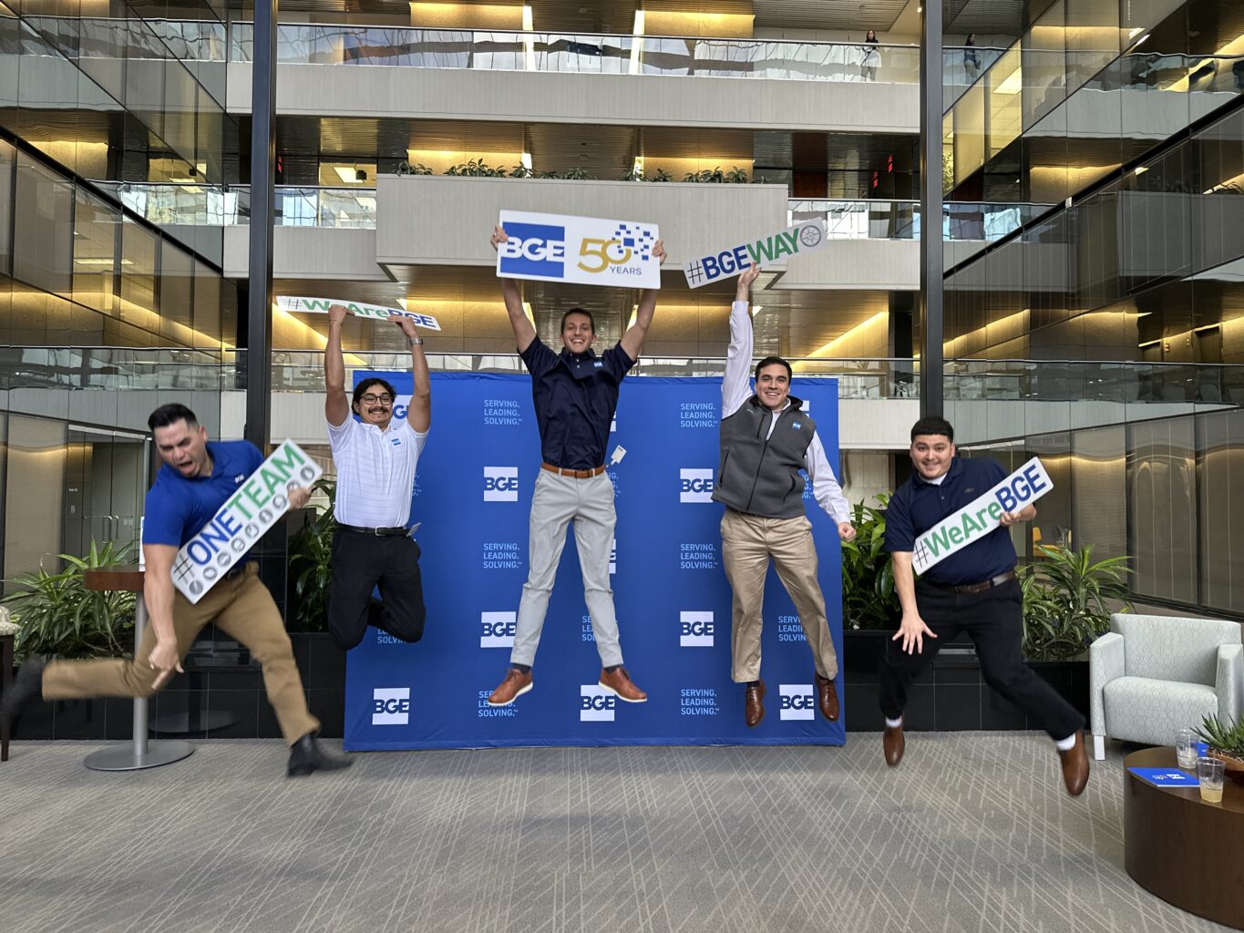 Five people jump in front of a blue BGE backdrop, holding signs that read “#ONEFAM,” “BGE 50 Years,” and “#WeAreBGE” to celebrate 50 years of excellence with BGE in an office lobby filled with plants and balconies.