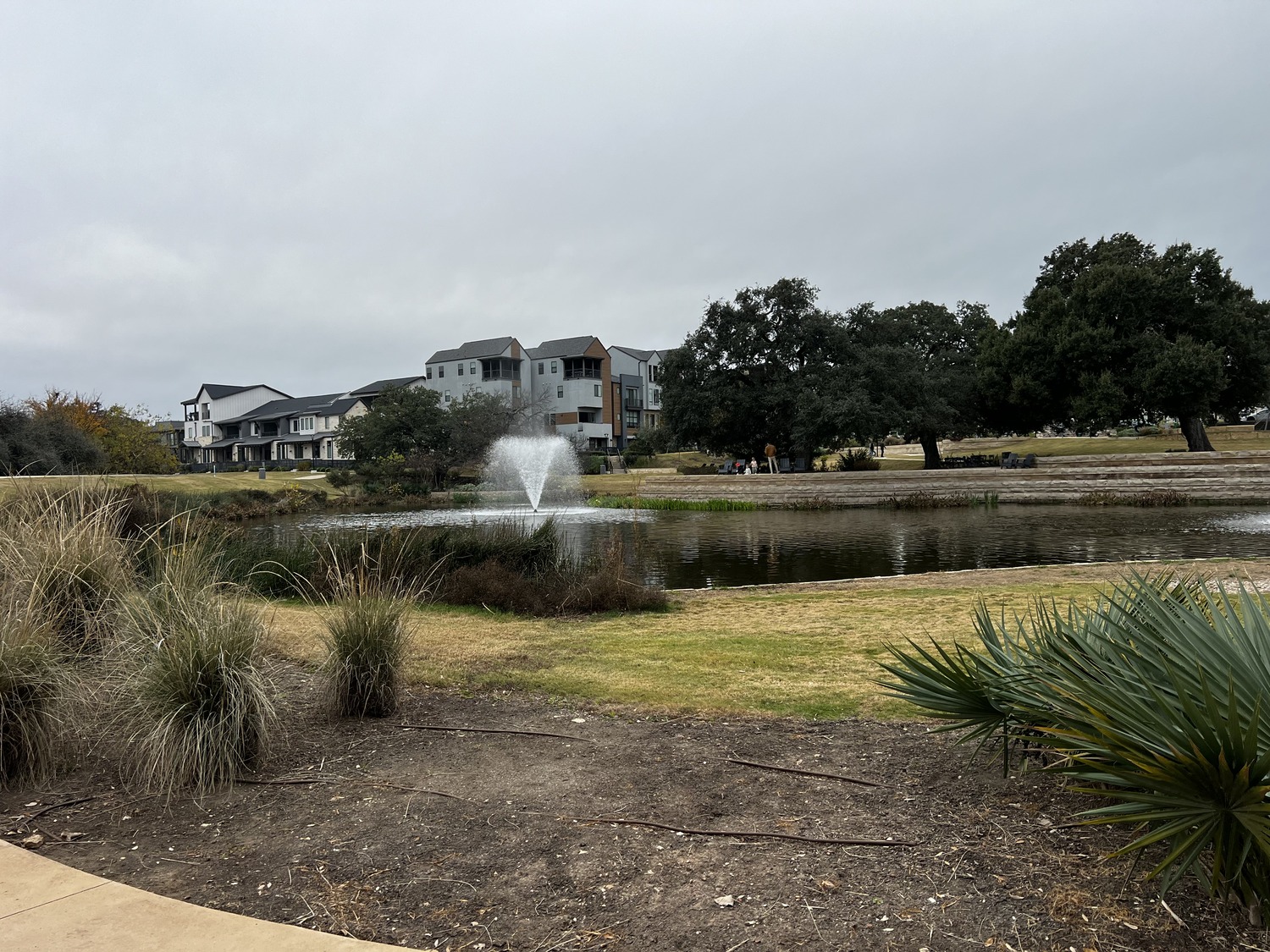 A pond with a water fountain is surrounded by grass, bushes, and trees at The Grove, with modern residential buildings in the background under a cloudy sky.