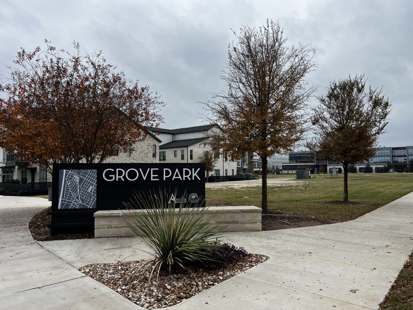 A black sign reading The Grove Park sits near a sidewalk, surrounded by trees with autumn leaves, grass, and buildings in the background under a cloudy sky.