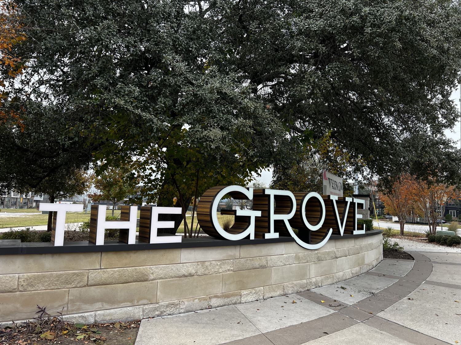 Large white letters spelling The Grove sit on a stone wall in front of trees with green and autumn-colored leaves, along a curved sidewalk in an outdoor park or community area, welcoming visitors to The Groveâs inviting atmosphere.