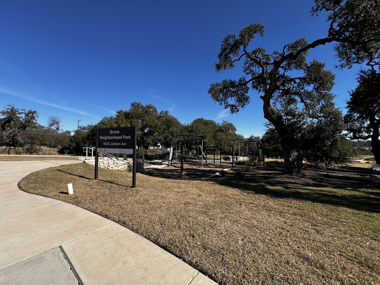A park with a sign reading The Grove Neighborhood Park, 4102 Jackson Ave, curved walkway, trees, playground equipment, and a clear blue sky.