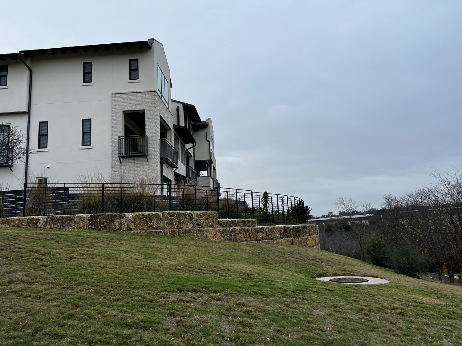 A modern white building with balconies sits on a grassy hill with stone retaining walls under a cloudy sky. To the right, trees and a road are visible in the distance, creating a tranquil setting that perfectly complements The Grove’s serene ambiance.
