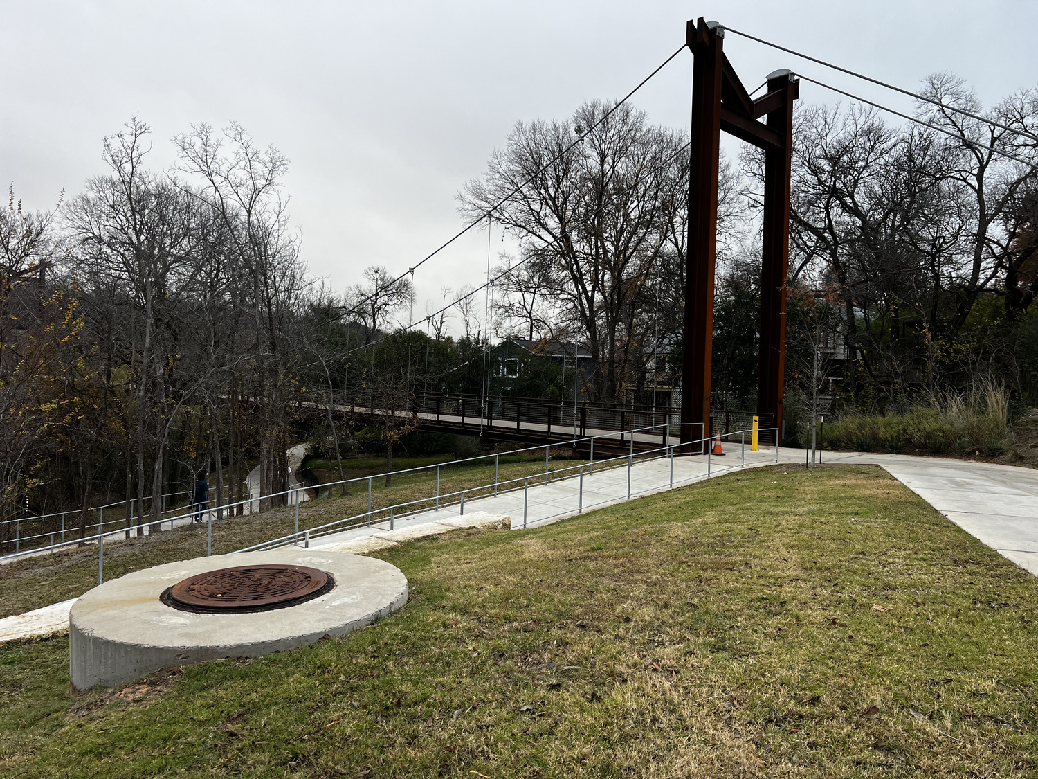 A modern pedestrian suspension bridge crosses a wooded area known as The Grove. In the foreground, there’s a concrete path, a grassy lawn, and a round metal manhole cover on a concrete base. Trees have sparse leaves, indicating late autumn or winter.