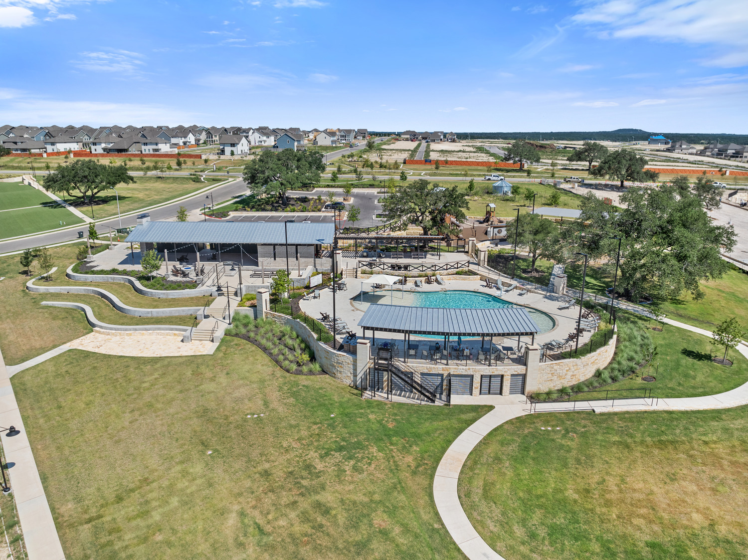 Aerial view of a modern neighborhood park in Lariat featuring a swimming pool, shaded seating area, walking paths, and landscaped green spaces, with houses and construction in the background under a blue sky.