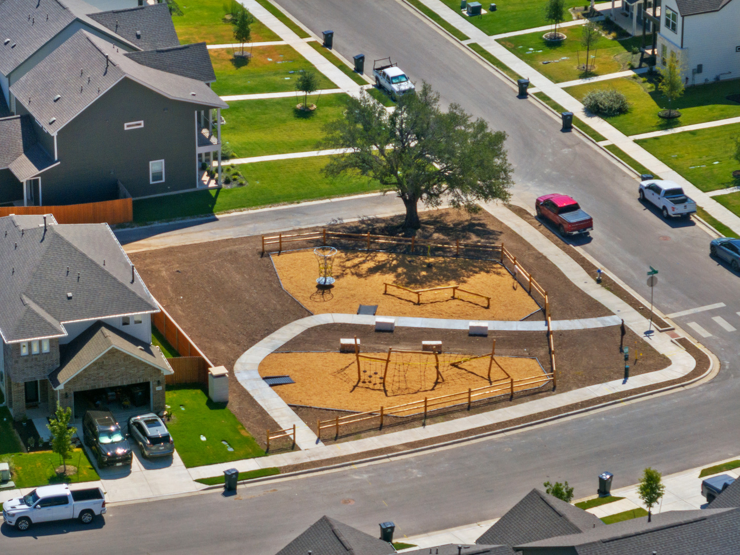 A small fenced playground with benches and play structures sits under a large tree in a residential neighborhood—where lariat-style ropes add an adventurous touch for kids, surrounded by houses, green lawns, and parked cars nearby.