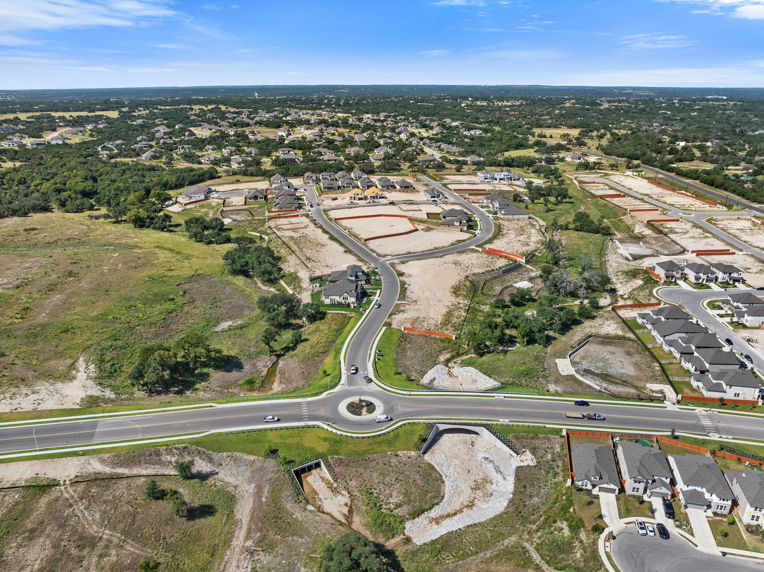 Aerial view of a suburban neighborhood under development, featuring a lariat-shaped roundabout, houses, construction areas, green spaces, and winding roads under a blue sky.