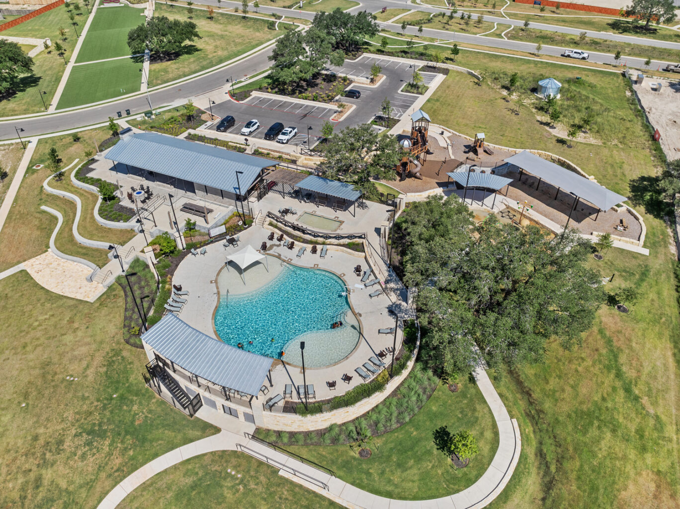 Aerial view of a public park featuring a swimming pool, shaded seating, playground, parking lot, green spaces, and winding lariat-shaped pathways under clear skies.