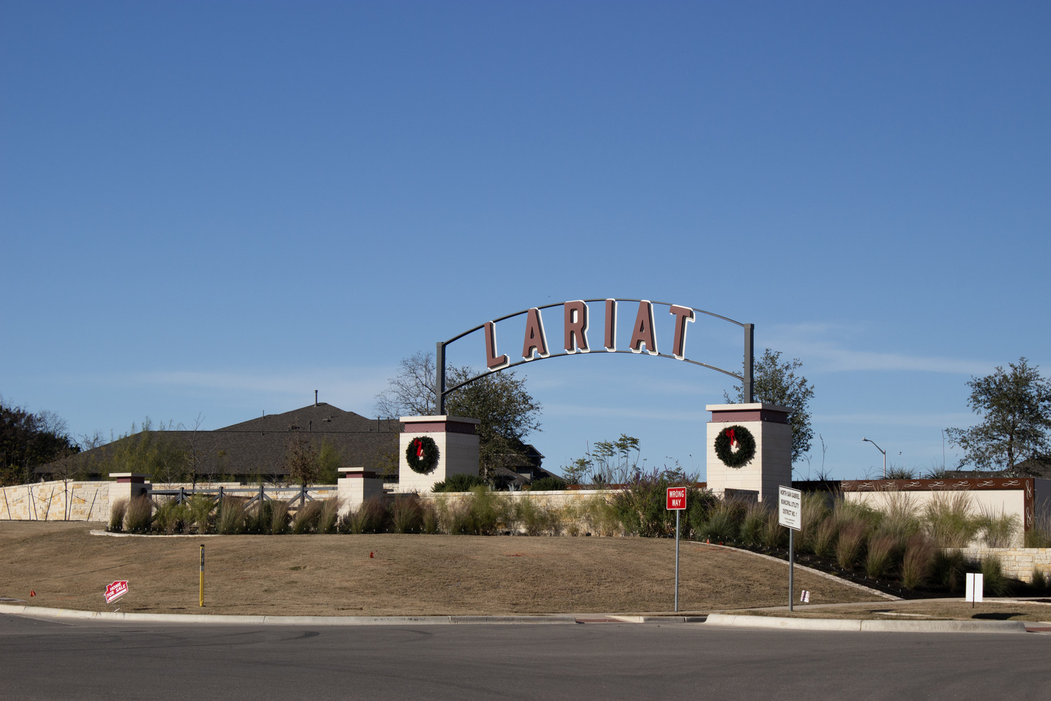 A large sign reading Lariat arches over a gated entrance decorated with holiday wreaths, welcoming visitors to the property, with a house, grass, and a clear blue sky in the background.