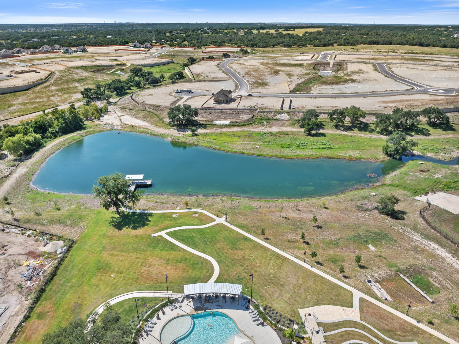 Aerial view of a pool and covered patio near a large pond surrounded by grass, trees, and dirt areas with construction and roads in the background, evoking the open charm of a Lariat landscape.