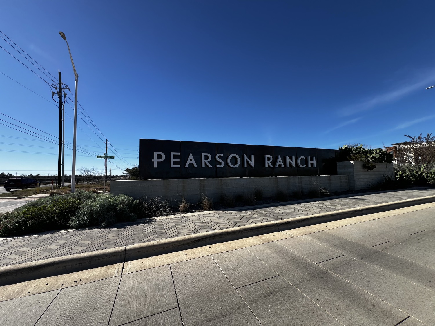 A large Pearson Ranch sign stands along a road, with a sidewalk, plants, and clear blue sky in the background.