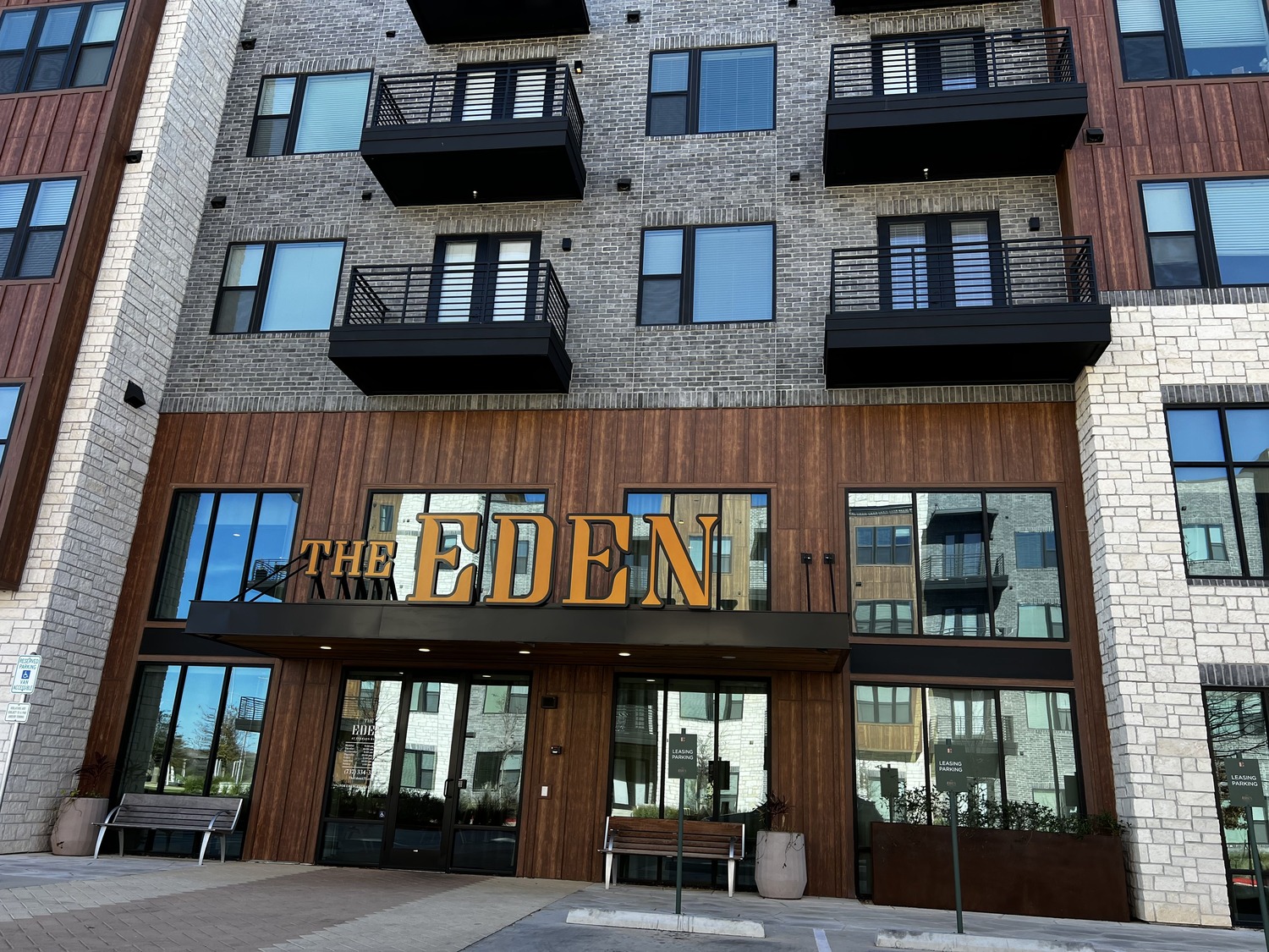 Modern apartment building exterior with large yellow letters spelling THE EDEN above the entrance, located in the vibrant Pearson Ranch area, featuring brick, wood, and stone facade, balconies, large windows, and benches by the doorway.