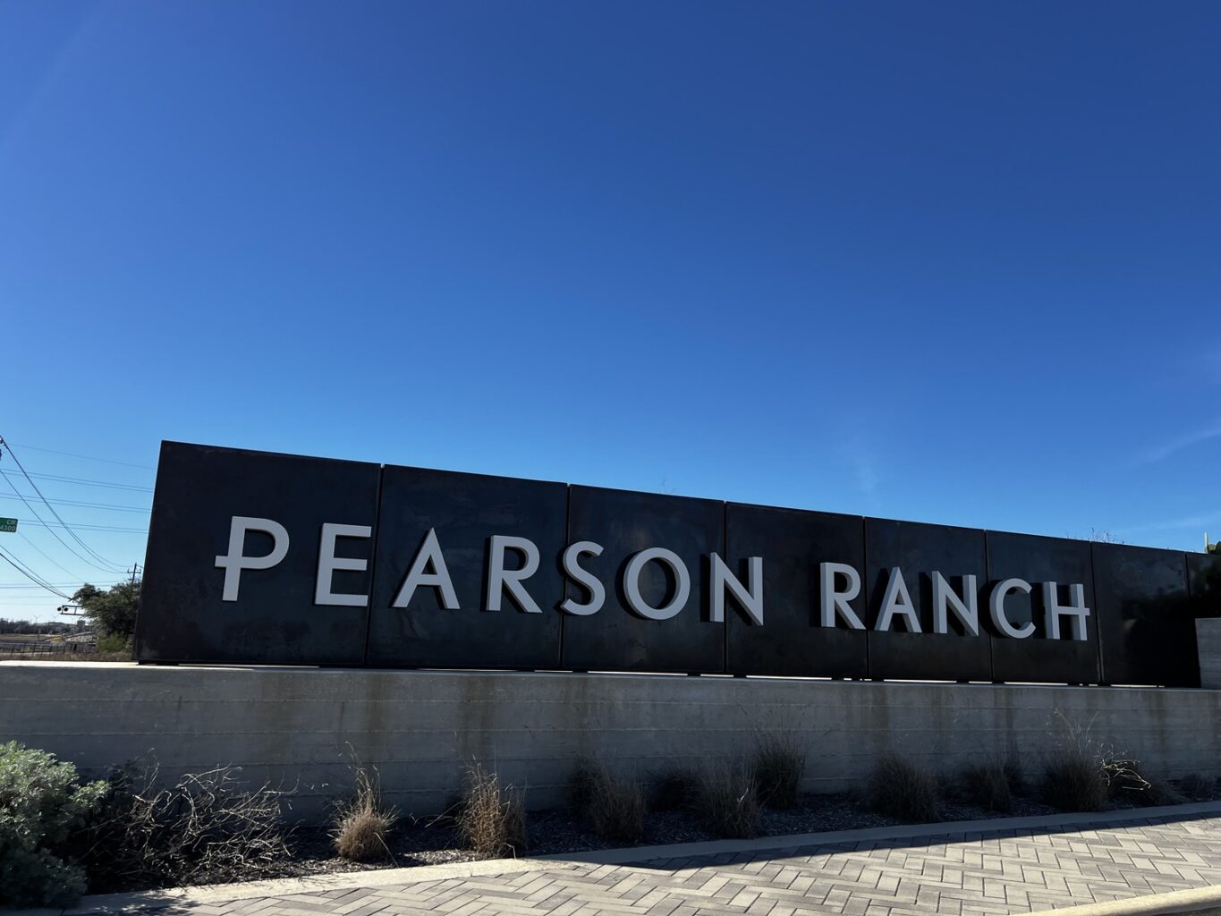 A large metal sign with bold white letters spells Pearson Ranch, prominently displayed on a low concrete wall. The sign stands surrounded by dry grasses, set against the backdrop of a clear blue sky.