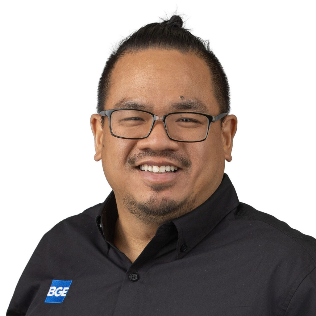 A smiling man with glasses, short hair in a top knot, and facial hair wears a black shirt with a blue BGE logo on the chest, standing against a plain white background—Steven Thai is captured in this friendly and approachable portrait.