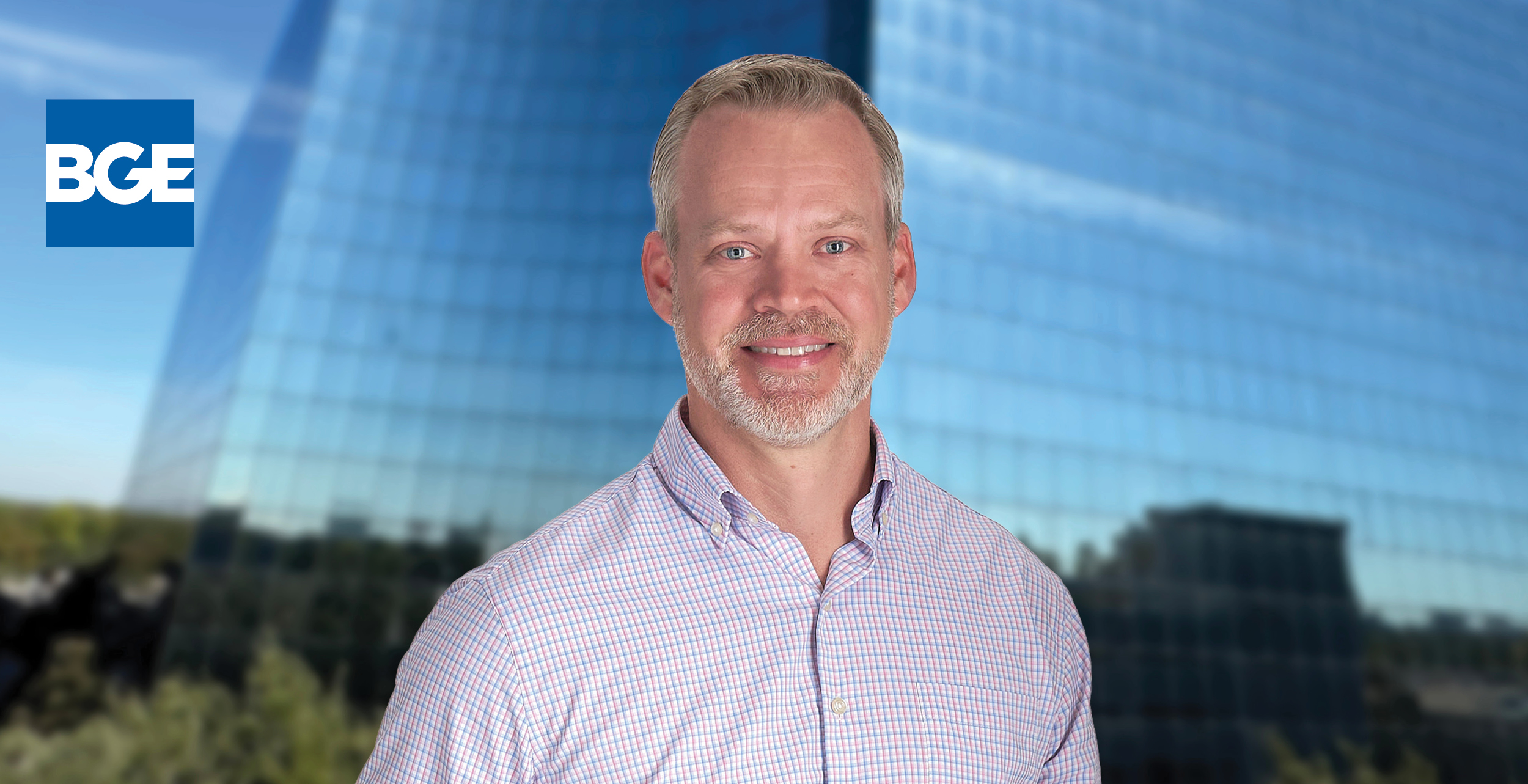 Jeff Safe, with short gray hair and a beard stands in front of a glass office building. He wears a light-colored, checked shirt.