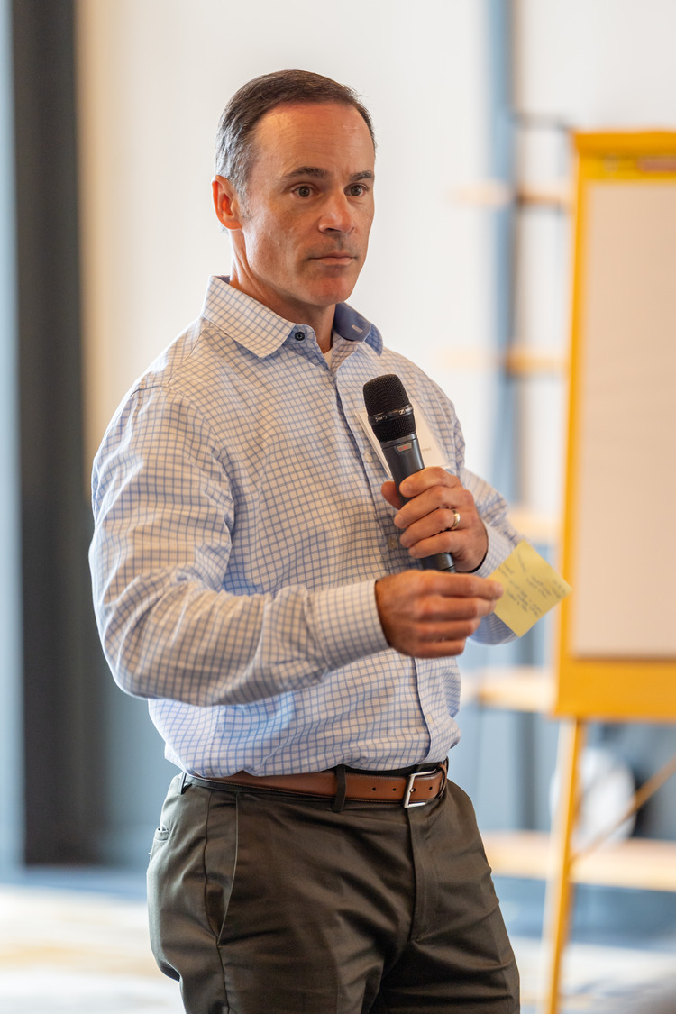 Michael Fitzgerald, dressed in a checkered shirt, holds a microphone and note cards while speaking in a bright room.
