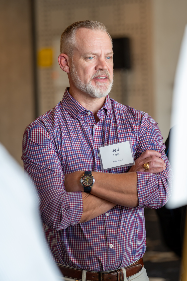 A man with gray hair and a beard, wearing a red checkered shirt and a name tag that says Jeff, stands with arms crossed and listens attentively indoors, reminiscent of the attentive demeanor.