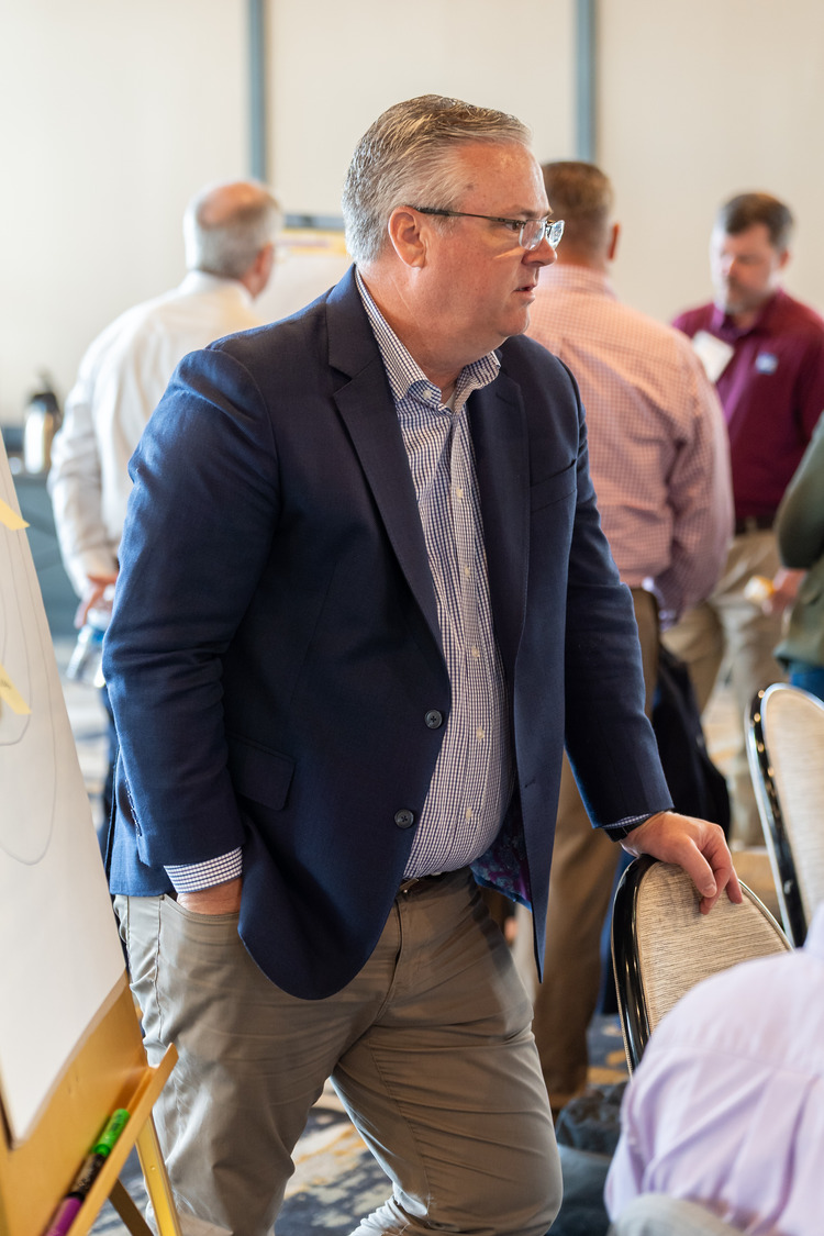 Chris Kuykendall, dressed in a sharp blue blazer and glasses, stands beside a chair, leaning on its back, while other people converse in the background at what appears to be a professional event or meeting.